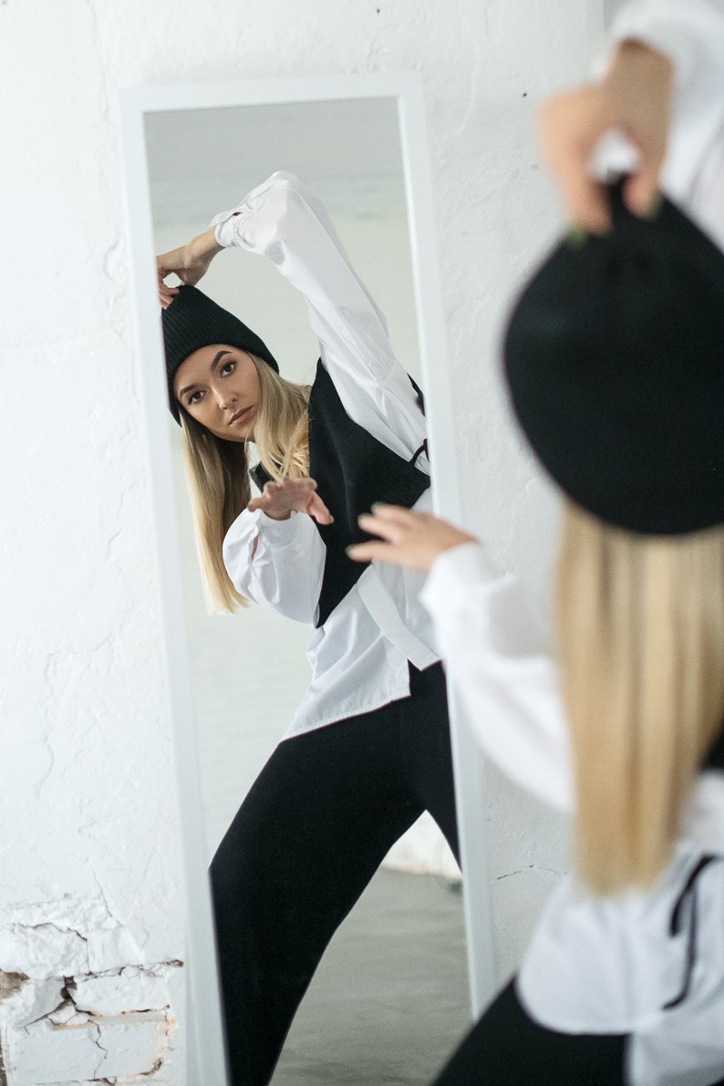 May include: A woman in a white shirt and black trousers poses in front of a mirror. She is wearing a black beanie and has her hair pulled back. She is looking at the camera.