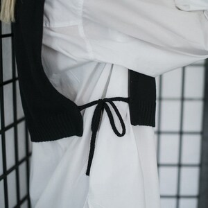 May include: A black knitted vest with a tie closure worn over a white button-down shirt.