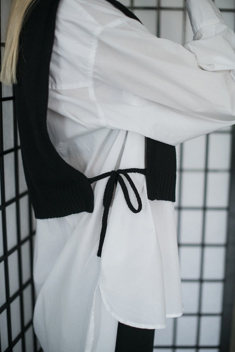May include: A black knitted vest with a tie closure worn over a white button-down shirt.