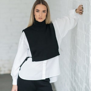 May include: A woman wearing a white button-down shirt layered over a black turtleneck sweater vest and black trousers.