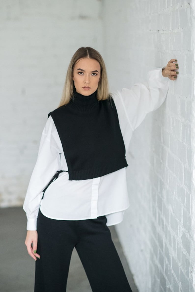 May include: A woman wearing a white button-down shirt layered over a black turtleneck sweater vest and black trousers.