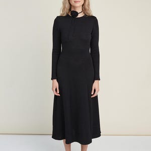 May include: A long-sleeved black midi dress with a round neckline and a flared skirt silhouette. The dress is paired with a black choker featuring a fabric flower accent and dark brown Mary Jane shoes with a buckle strap closure.