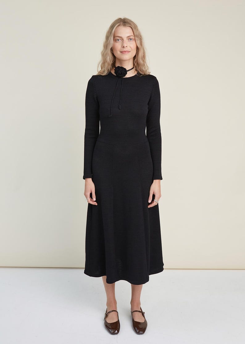 May include: A long-sleeved black midi dress with a round neckline and a flared skirt silhouette. The dress is paired with a black choker featuring a fabric flower accent and dark brown Mary Jane shoes with a buckle strap closure.