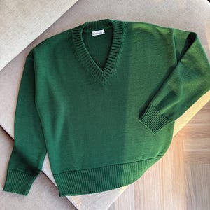 Puede incluir: Un suéter verde con cuello en V con puños y dobladillo acanalados. El suéter está hecho de un material de punto suave y tiene una etiqueta que dice "mouala". El suéter se muestra sobre una superficie de color neutro.