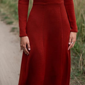 May include: A long-sleeved, rust-red knit dress featuring a fitted bodice, a defined waistline, and a flared skirt. The garment is constructed with a fine, uniform knit texture and includes vertical seam detailing that extends from the waist.