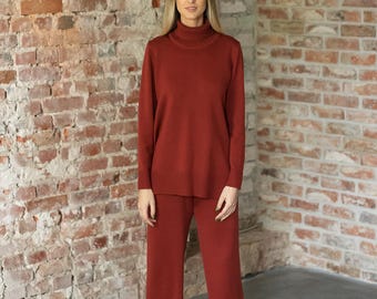 May include: A rust-colored two-piece set consisting of a long-sleeved turtleneck sweater with ribbed cuffs and hem, paired with matching wide-leg trousers. The knit garment features a solid, deep terracotta tone throughout the entire outfit.