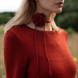 May include: A dark red long-sleeved knit sweater paired with a matching dark red crocheted rose choker. The choker features a central floral design with two thin dangling cords that end in small, rounded, teardrop-shaped decorative accents.