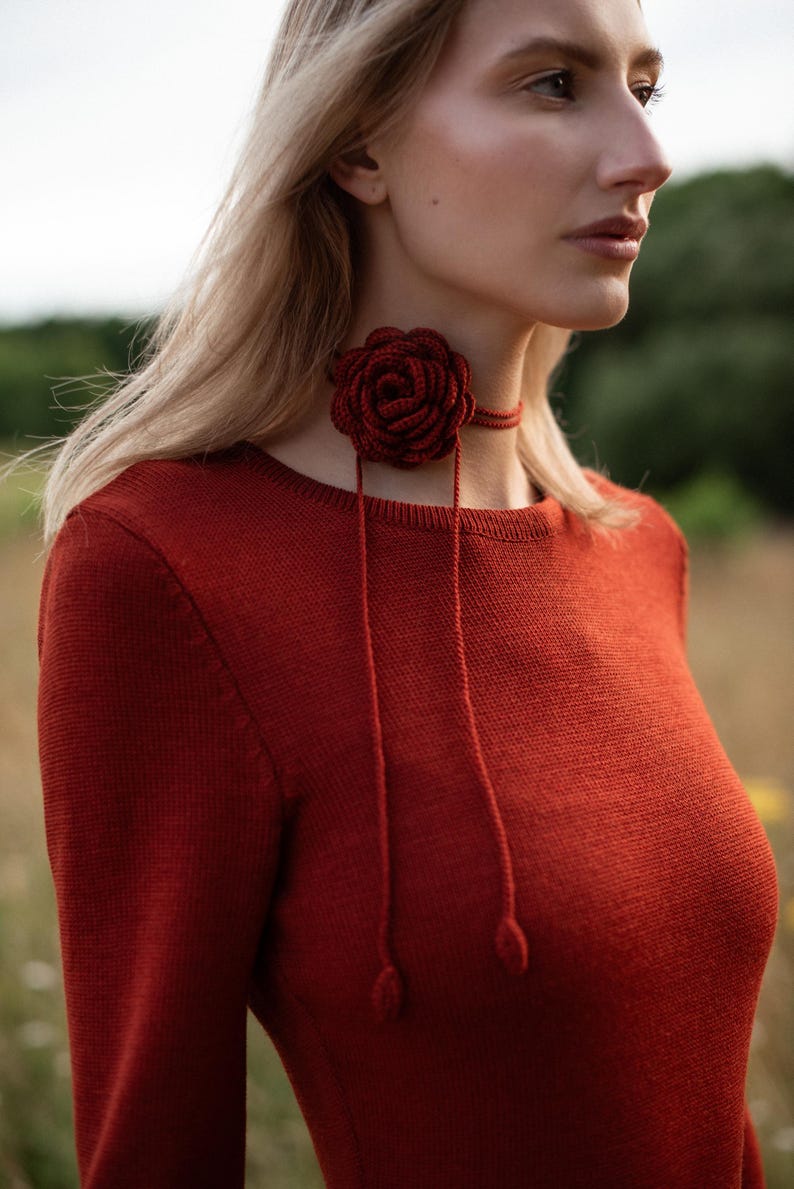May include: A dark red long-sleeved knit sweater paired with a matching dark red crocheted rose choker. The choker features a central floral design with two thin dangling cords that end in small, rounded, teardrop-shaped decorative accents.