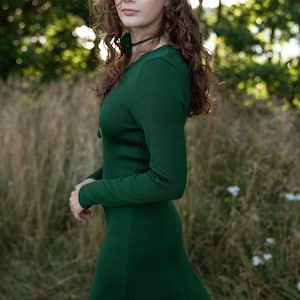 May include: A long-sleeved, dark green knit dress featuring a fitted bodice, a round neckline, and a flared skirt. The garment is constructed from a solid-colored, textured fabric with a subtle ribbed pattern throughout the material.