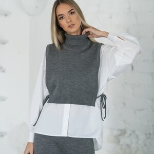 May include: A woman wearing a grey turtleneck sweater vest over a white button-down shirt and matching grey trousers.