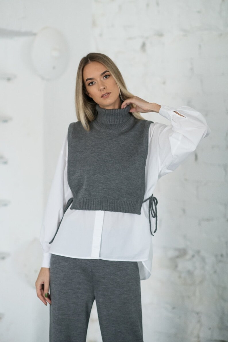 May include: A woman wearing a grey turtleneck sweater vest over a white button-down shirt and matching grey trousers.