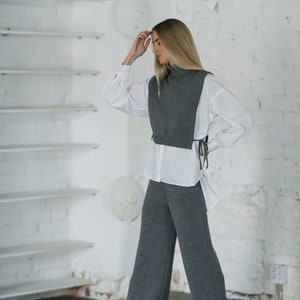 May include: A woman wearing a grey turtleneck sweater vest with a white button-down shirt and grey wide-leg trousers.