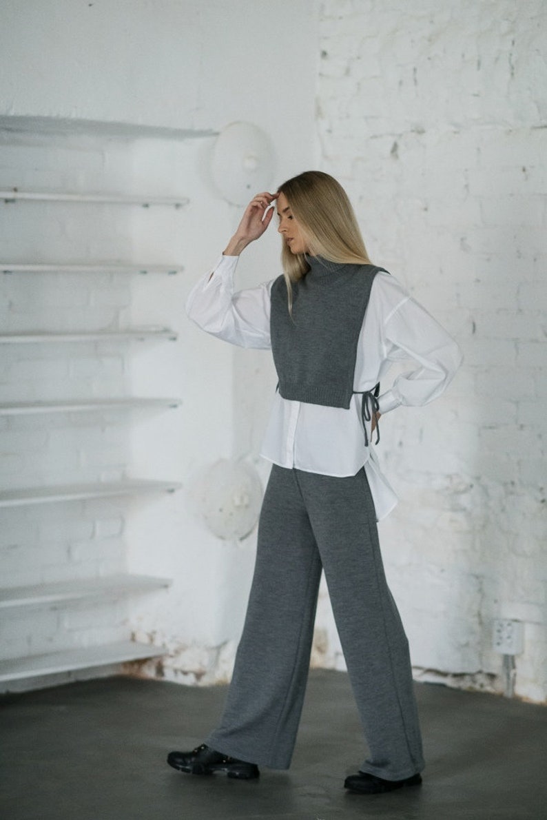 May include: A woman wearing a grey turtleneck sweater vest with a white button-down shirt and grey wide-leg trousers.