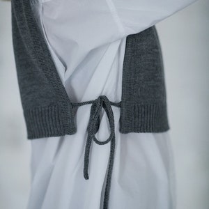 May include: A grey knitted vest with a tie closure worn over a white button-down shirt.