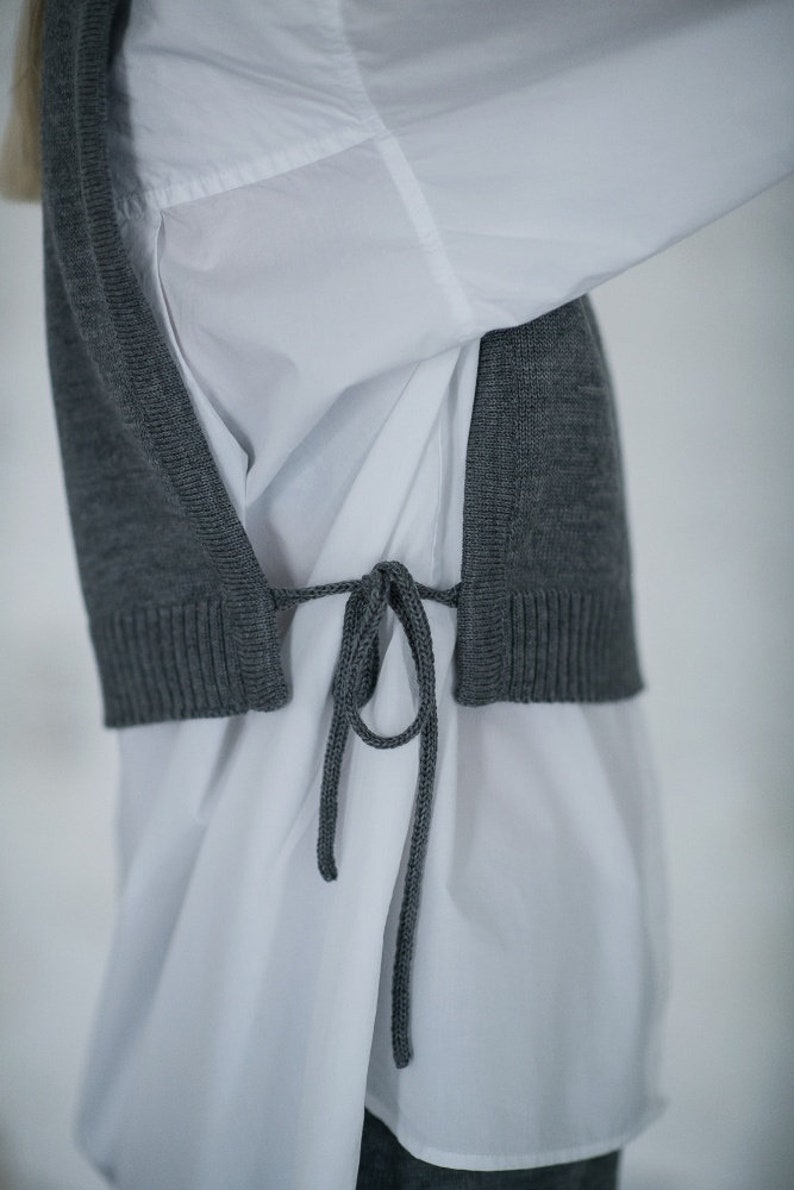 May include: A grey knitted vest with a tie closure worn over a white button-down shirt.