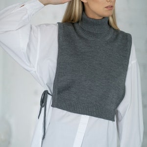 May include: A grey turtleneck sweater vest worn over a white button-down shirt. The vest has a relaxed fit and is cropped at the waist. The shirt has long sleeves and is tucked into the vest.
