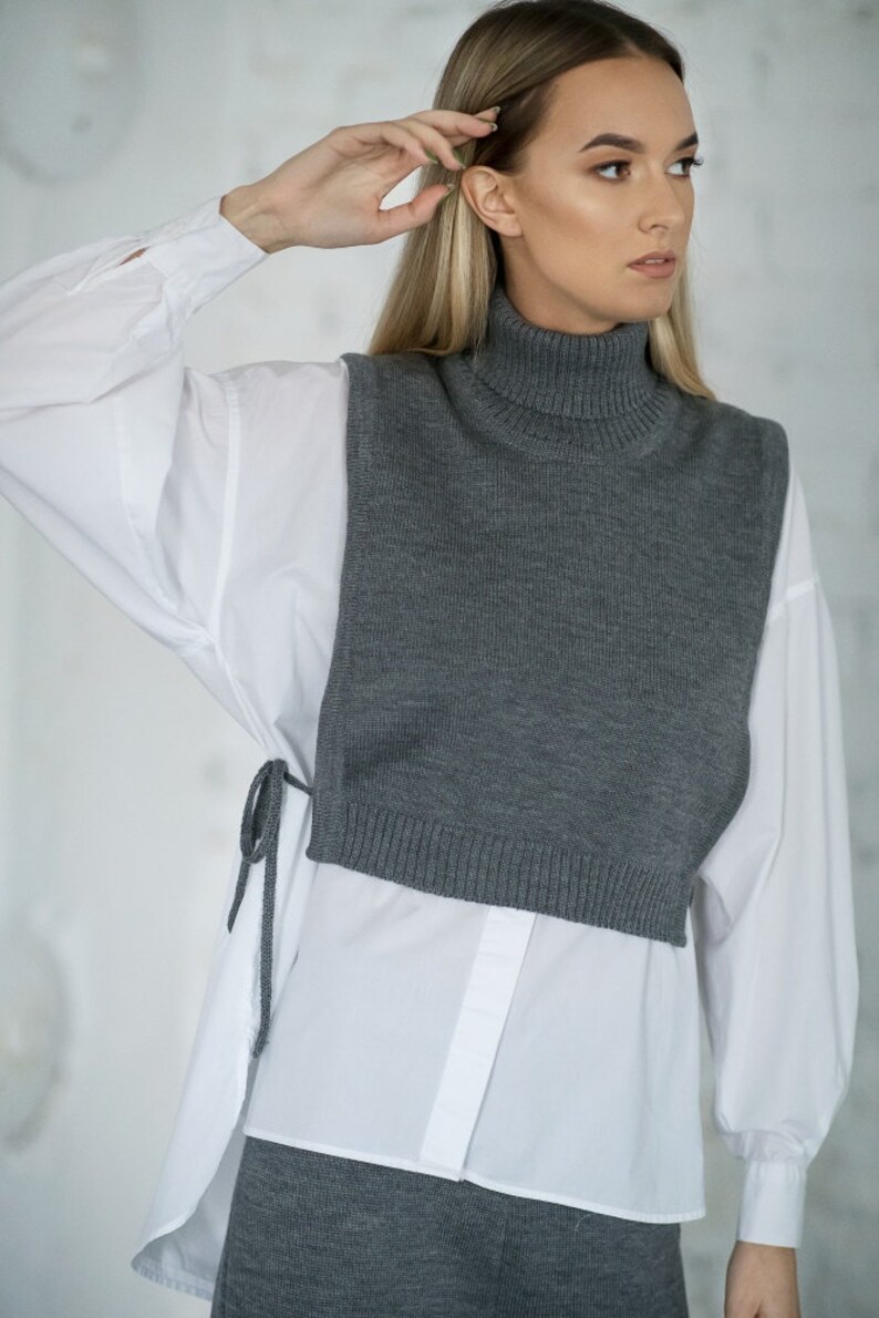 May include: A grey turtleneck sweater vest worn over a white button-down shirt. The vest has a relaxed fit and is cropped at the waist. The shirt has long sleeves and is tucked into the vest.