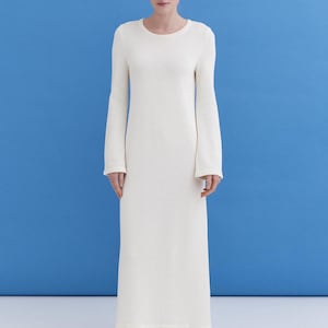 Peut inclure: Une longue robe blanc cassé à manches longues et col rond. La robe a un design simple et élégant et descend jusqu'aux chevilles. La modèle porte des ballerines bleues. Le fond est bleu uni.