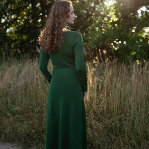 May include: A long-sleeved, ankle-length dress in a solid dark green color. The garment features a fitted bodice that transitions into a flared skirt, with a simple round neckline and a smooth, knit-like texture visible from the back view.