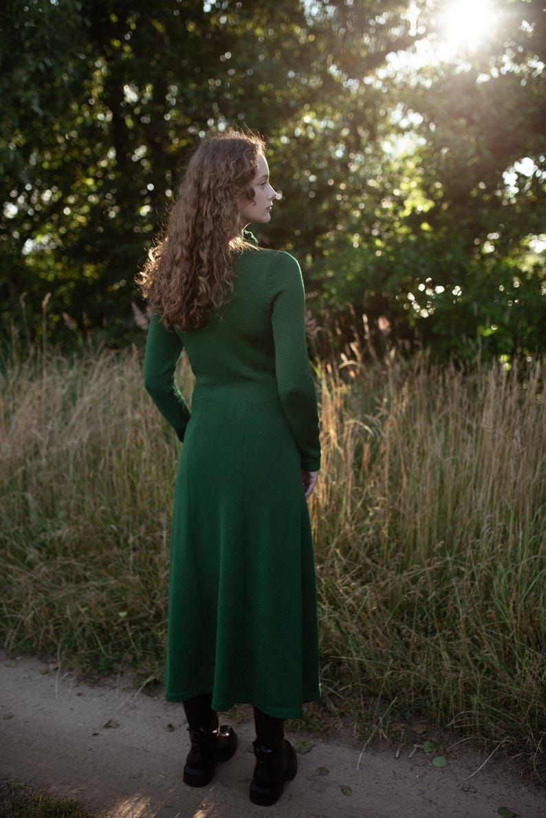 May include: A long-sleeved, ankle-length dress in a solid dark green color. The garment features a fitted bodice that transitions into a flared skirt, with a simple round neckline and a smooth, knit-like texture visible from the back view.
