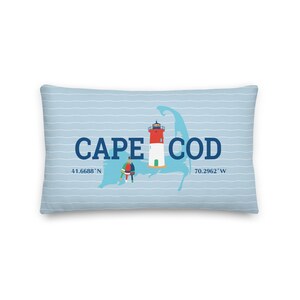 May include: A blue and white striped pillow with a white outline of Cape Cod, a red and white lighthouse, and the text "CAPE COD" in blue. The coordinates "41.6688' N" and "70.2962' W" are also included in blue.