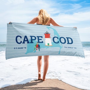 May include: A light blue beach towel with horizontal stripes and the words "CAPE COD" in navy blue. The towel features a map outline of Cape Cod, a lighthouse, and coordinates. The towel is held up by a person on a beach.