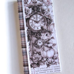 May include: A rectangular bookmark featuring a brown and tan plaid fabric spine. The main panel displays a sepia-toned illustration of a rabbit wearing a top hat and vest, standing beside a large clock, surrounded by roses and French text.