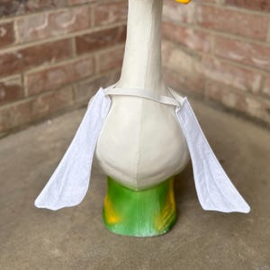 May include: A whimsical, decorative goose statue. The goose has a white body, a yellow beak, and black eyes. It features white wings and a green and yellow base. The statue is designed to stand upright and is suitable for outdoor or indoor display.