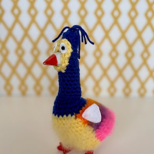 May include: A handmade crochet duck with a blue head, yellow body, and colorful tail feathers. The duck has a red beak and feet. The background is a white surface with a gold diamond pattern.
