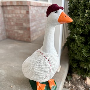 May include: A white goose statue wearing a burgundy baseball cap and a white crocheted baseball-themed sweater. The goose has an orange beak and feet, standing on a green base. The statue is displayed outdoors.