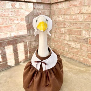 May include: A goose costume featuring a white headpiece with a yellow beak and black eyes, and a brown dress with a white collar and a brown bow. The costume is designed to fit a goose.