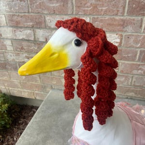 Könnte beinhalten: Eine weiße Gans, die eine rote gehäkelte Perücke mit langen, lockigen Strähnen trägt. Die Gans trägt auch ein pinkfarbenes Paillettenkleid.