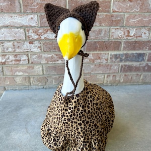 May include: A goose wearing a brown crocheted hat with ear flaps and a leopard print costume. The goose has a white head with a yellow beak and a brown string around its neck. The costume has a ruffled hem.