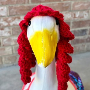 May include: A white goose head with a yellow beak and black eyes, wearing a red crocheted wig and a pink and red crocheted bikini top. The background is a brick wall.