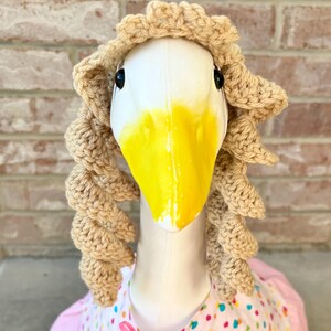 May include: A goose head with a white face and yellow beak, wearing a tan crocheted wig. The goose is dressed in a pink and white dress with colorful polka dots and a pink bow.