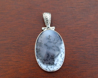 Puede incluir: Un colgante de plata con una piedra de ágata dendrítica blanca y negra. La piedra tiene forma ovalada y tiene un aro de plata.