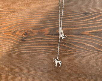 May include: Silver-toned necklace featuring a horse pendant and a stirrup charm. The chain has a delicate, twisted design. The horse pendant is a detailed miniature sculpture. The necklace is displayed against a wooden background.