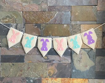 May include: A felt banner with seven pennants featuring bunny silhouettes in pink, purple, and blue. Each pennant has a white button in the center.