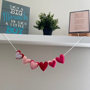 K&ouml;nnte beinhalten: Eine wei&szlig;e Schnur mit einer Girlande aus Filzherzen in Rosa, Rot und Wei&szlig;, die an einem wei&szlig;en Regal h&auml;ngt. Das Regal hat auch ein graues Schild mit dem Text "Such a Big Miracle in Such a Little Boy" und eine kleine Topfpflanze. Ein gerahmtes Bild mit spanischem Text befindet sich ebenfalls auf dem Regal.