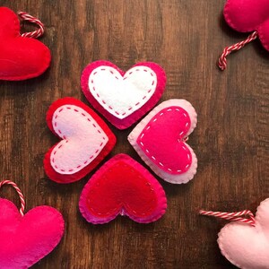 Op de afbeelding: Vijf vilten harten in verschillende tinten roze en rood, gerangschikt in een cirkel op een bruine houten achtergrond. De harten zijn gestikt met wit garen.