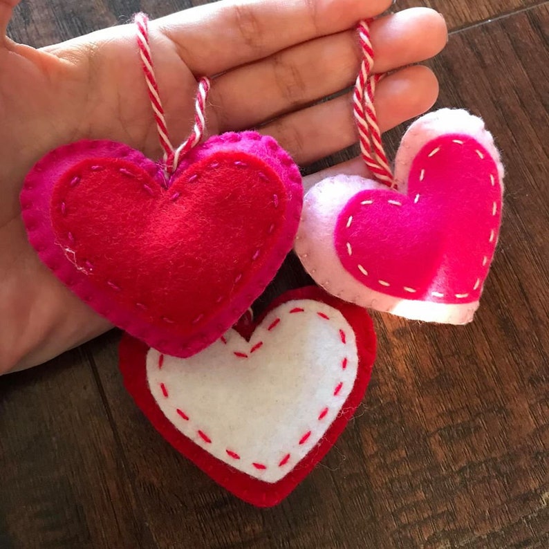 Op de afbeelding: Drie vilten hartvormige ornamenten in roze, rood en wit met witte stiksels rond de randen. De harten hangen aan een wit touwtje.