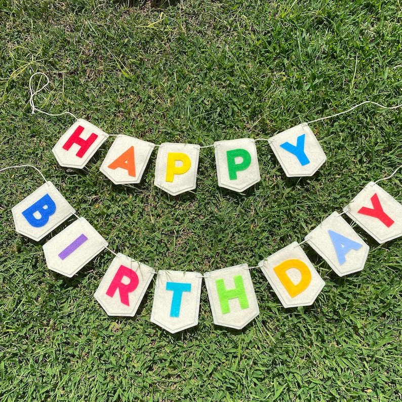 Pu&ograve; includere: Un festoso striscione "Happy Birthday" con lettere colorate su bandierine color crema. Le lettere sono rosse, arancioni, gialle, verdi, blu, viola e azzurre. Lo striscione &egrave; infilato su uno spago bianco su uno sfondo erboso verde.