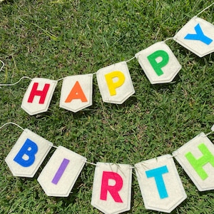 Pu&ograve; includere: Un festoso striscione con la scritta "HAPPY BIRTHDAY" in lettere di feltro colorate. Ogni lettera &egrave; di un colore diverso, attaccata a bandierine bianco sporco. Lo striscione &egrave; infilato su uno spago bianco, perfetto per una festa di compleanno.