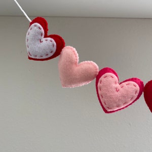 K&ouml;nnte beinhalten: Eine Girlande aus Filzherzen in Rosa- und Rott&ouml;nen, die mit wei&szlig;em Garn zusammengef&uuml;gt sind. Die Herzen haben ein schlichtes gesticktes Design.