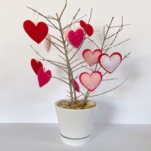 Op de afbeelding: Een witte keramische pot met kleine stenen erin, versierd met vilten harten in roze en rode tinten die aan kale takken hangen.
