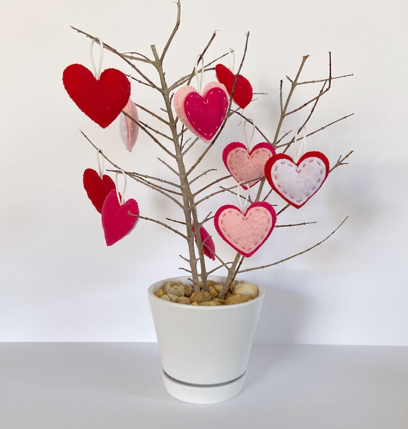 Op de afbeelding: Een witte keramische pot met kleine stenen erin, versierd met vilten harten in roze en rode tinten die aan kale takken hangen.