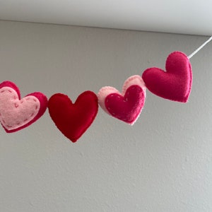 K&ouml;nnte beinhalten: Eine Girlande aus Filzherzen in Rosa und Rot, die mit wei&szlig;em Garn zusammengef&uuml;gt sind. Die Herzen sind mit einem einfachen Stich gen&auml;ht.