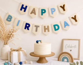 May include: A Happy Birthday felt banner with cream-colored pennants and multi-colored letters in blue, teal, and brown. Below, a white frosted cake sits on a wooden stand, accompanied by a gift box, a rainbow decor piece, and a framed sign.