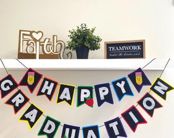 Könnte beinhalten: Ein farbenfrohes Abschlussbanner mit den Worten "HAPPY GRADUATION" in weißen Buchstaben auf schwarzen Wimpeln. Das Banner enthält Bleistift- und Apfelmotive. Ein Regal darüber zeigt ein "Faith"-Schild, eine Pflanze und ein "TEAMWORK"-Schild.