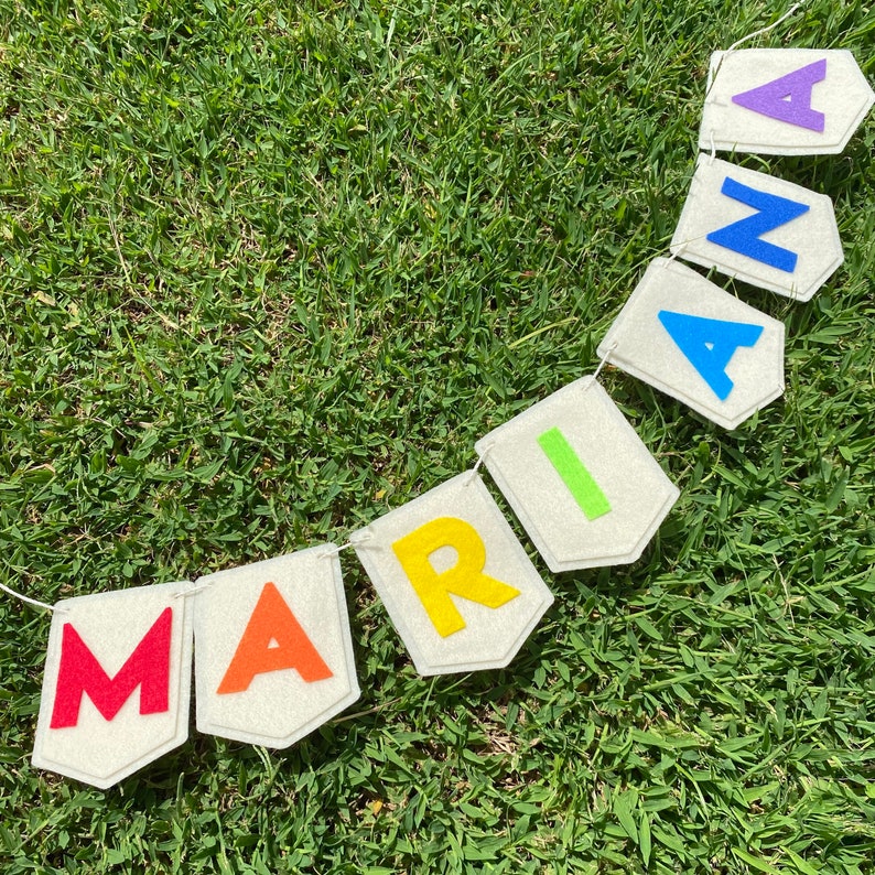 Pu&ograve; includere: Un banner fatto a mano che riporta il nome "MARIANA" in lettere di feltro colorate. Ogni lettera &egrave; di un colore diverso, attaccata a un gagliardetto di feltro di colore chiaro. Lo striscione &egrave; infilato su uno spago bianco sull'erba verde.